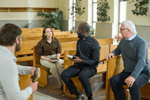 alt=’A small group seated in church pews, discussing while holding religious texts.’, aria-hidden=’true’