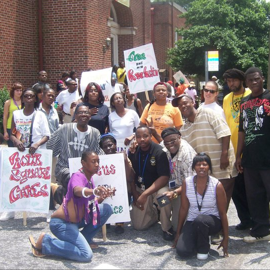 alt=’A diverse group of men, women, and children outdoors, holding signs’, aria-hidden=’true’